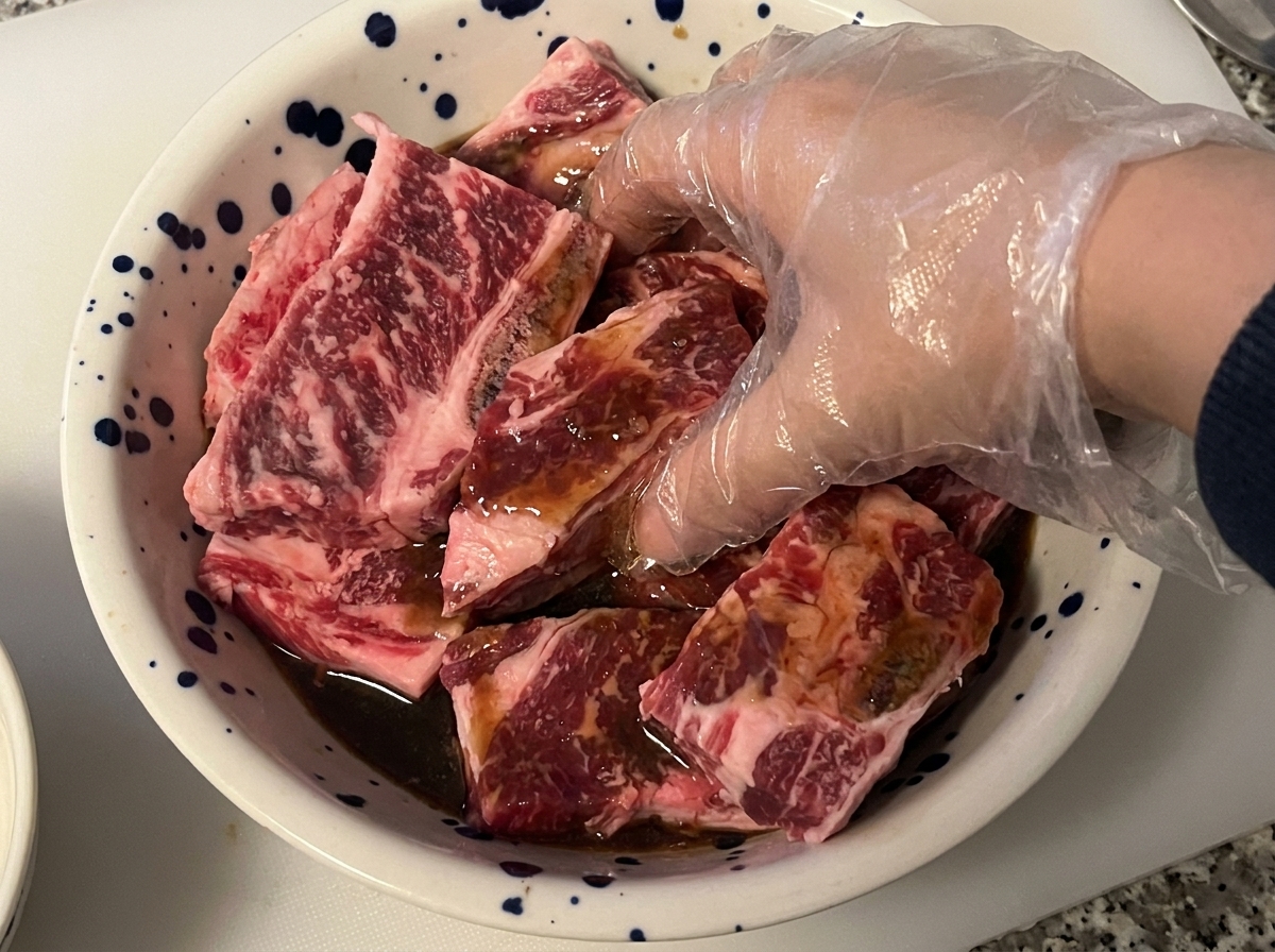 Eine handschuhte Hand mischt und massiert rohe Rinder-Kurzrippen mit dunkler Marinade in einer Schüssel.