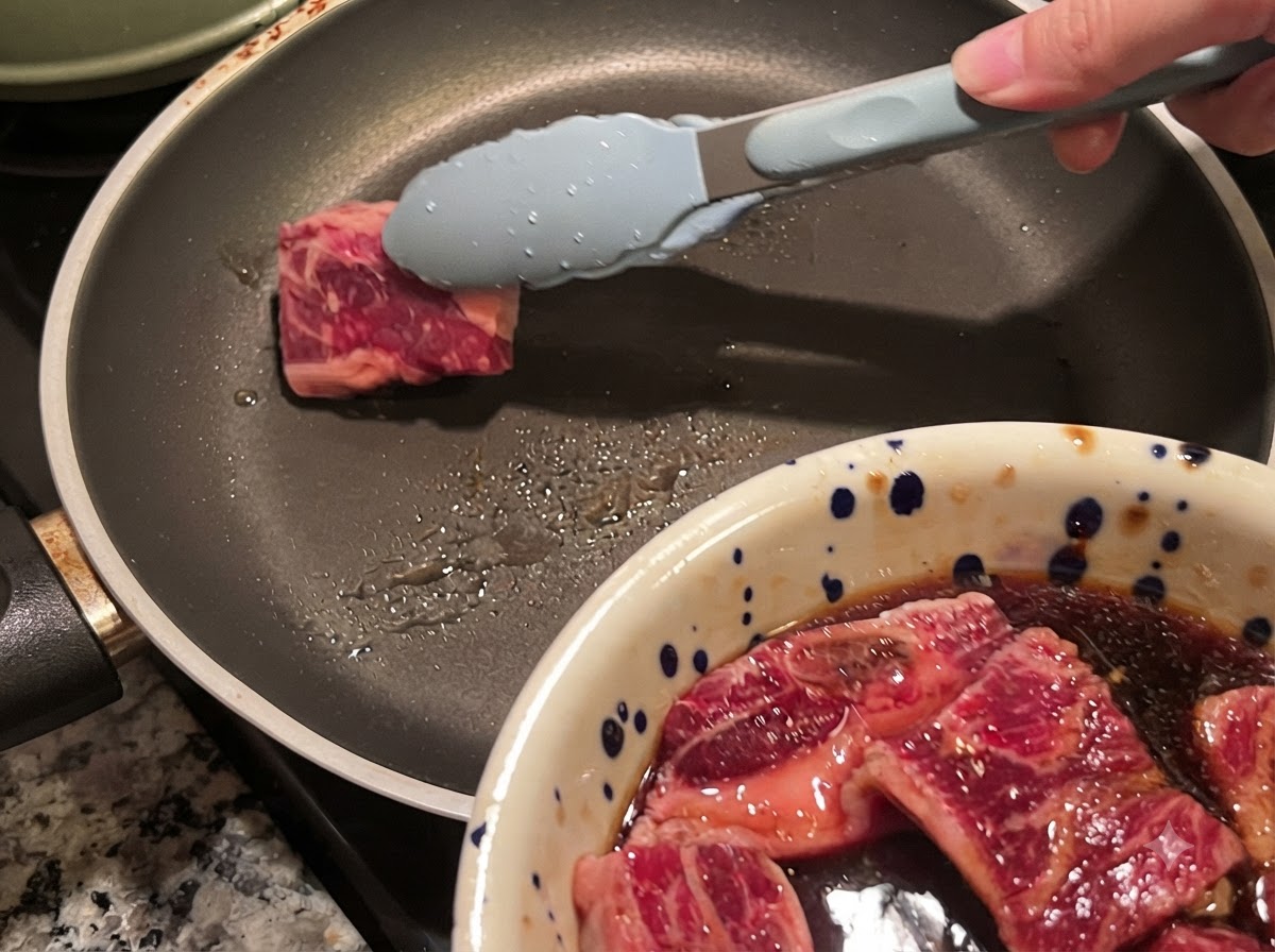Eine Zange legt ein Stück mariniertes Rinder-Kurzrippenfleisch in eine heiße Pfanne.