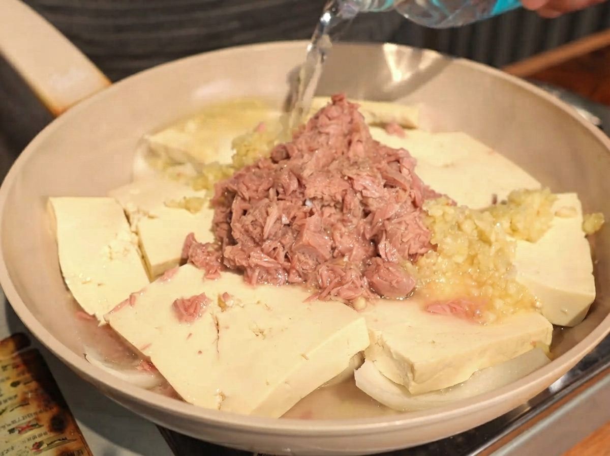 Klares Wasser wird aus einer Flasche in eine Pfanne mit Tofu, Zwiebeln, Knoblauch und Thunfisch gegossen.