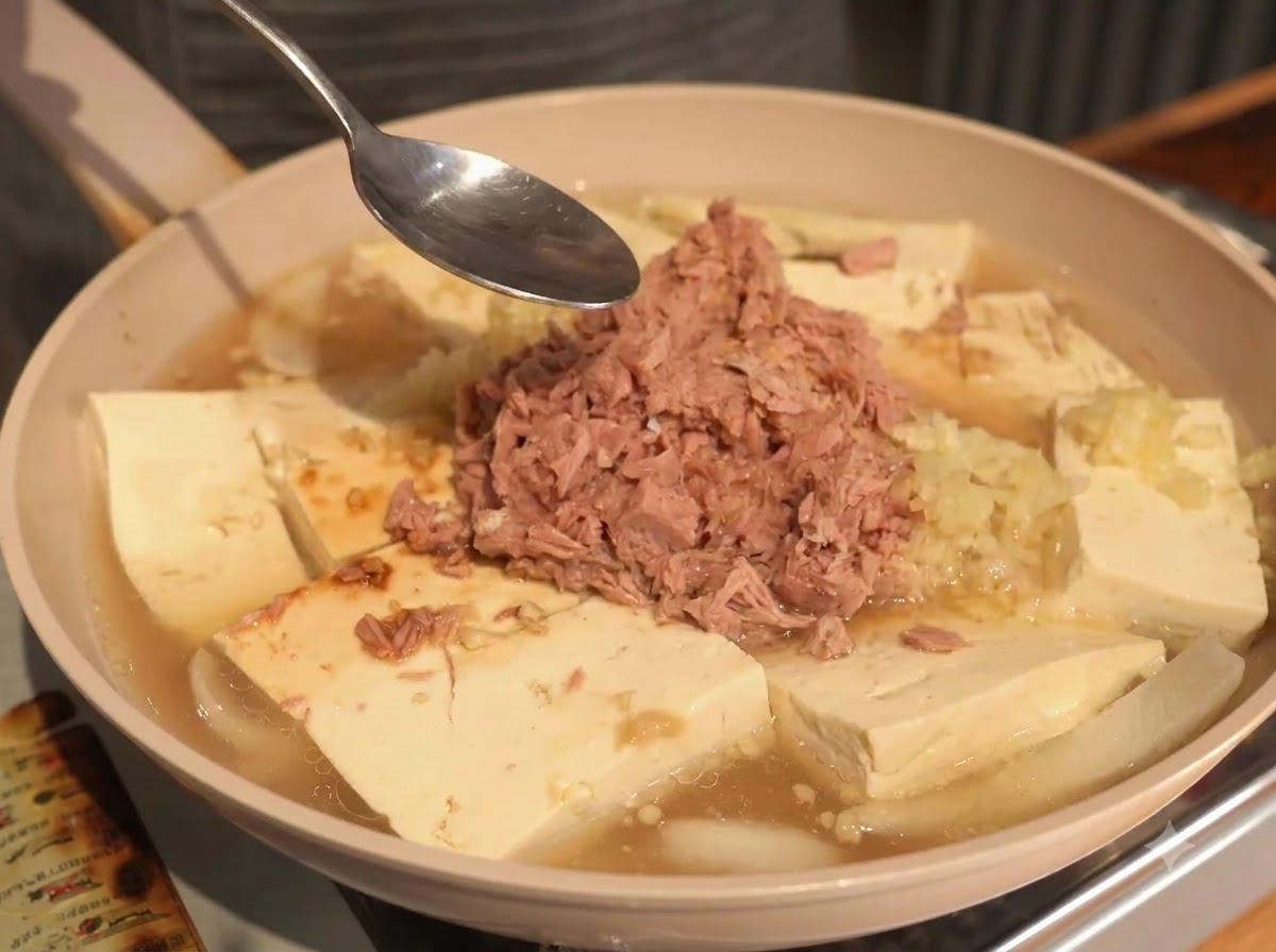 Ein Löffel über der Pfanne mit Thunfisch, Tofu und Flüssigkeit während des Würzevorgangs.