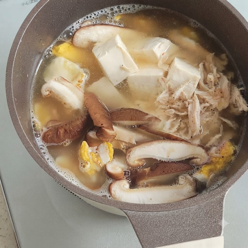 Ein kleiner Haufen getrockneter Garnelen liegt auf köchelndem Tofu, Pilzen und Ei in einem Topf mit Brühe.