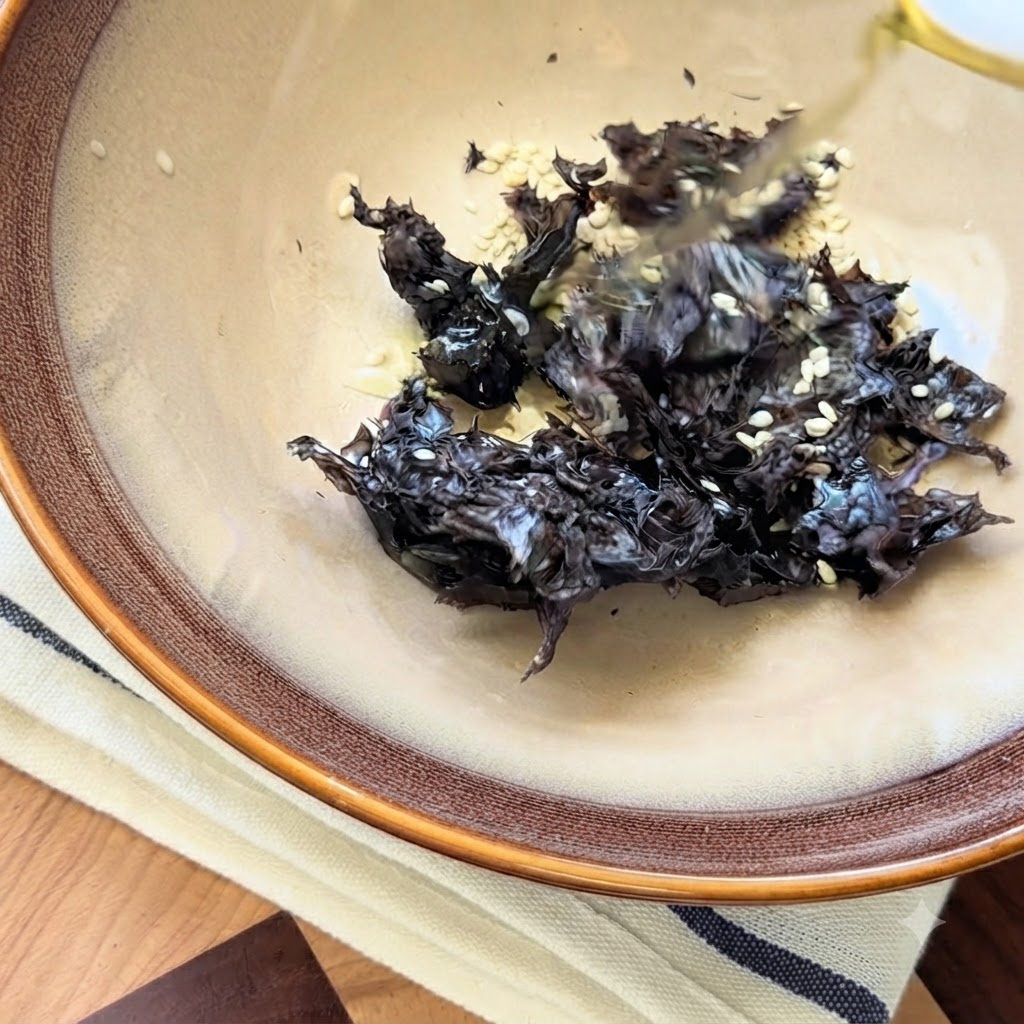Sichuan-Pfefferöl wird über getrocknete Algen und weiße Sesamsamen in einer Keramikschüssel geträufelt.