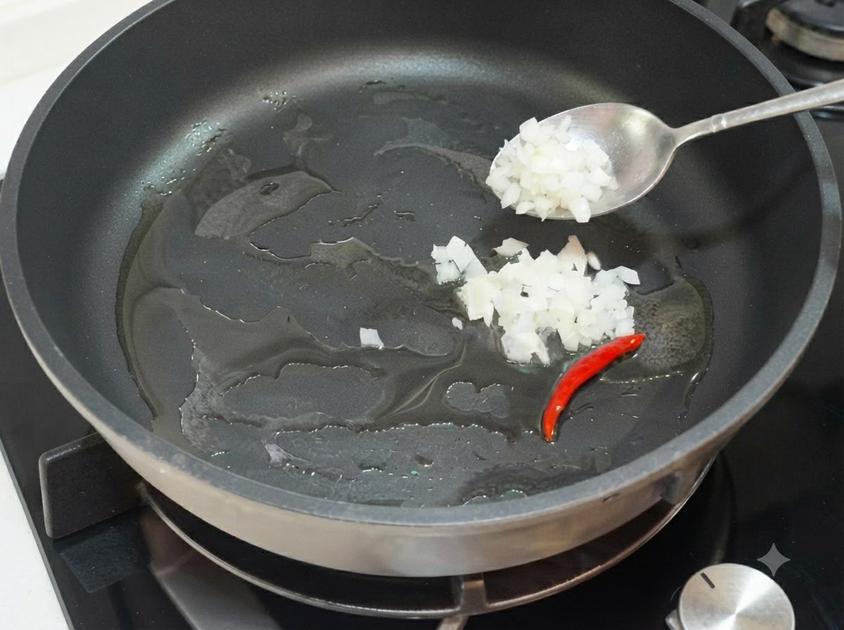 Eine Bratpfanne auf dem Herd mit Speiseöl, einer roten Chilischote und gewürfelten Zwiebelstücken, die mit einem Löffel hinzugefügt werden.
