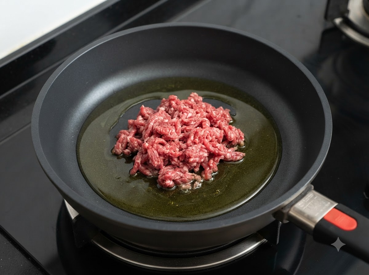 Rohes Hackfleisch, das in der Mitte einer Bratpfanne aufgehäuft ist, in der sich eine Pfütze Olivenöl auf einem Herd befindet.