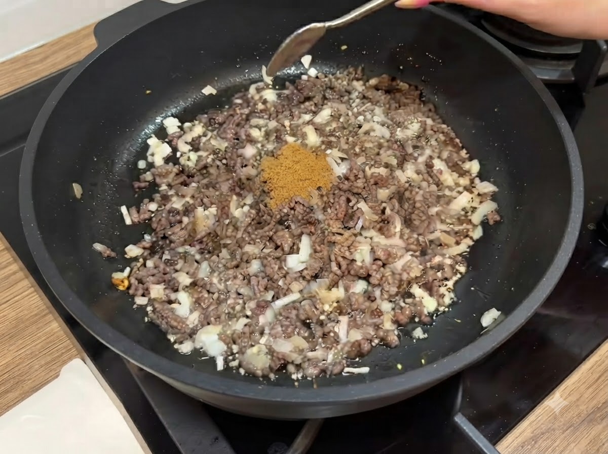Ein Löffel braunes Kreuzkümmelpulver wird in eine Bratpfanne gegeben, die mit gewürztem, gekochtem Hackfleisch und gehackten Zwiebeln gefüllt ist.