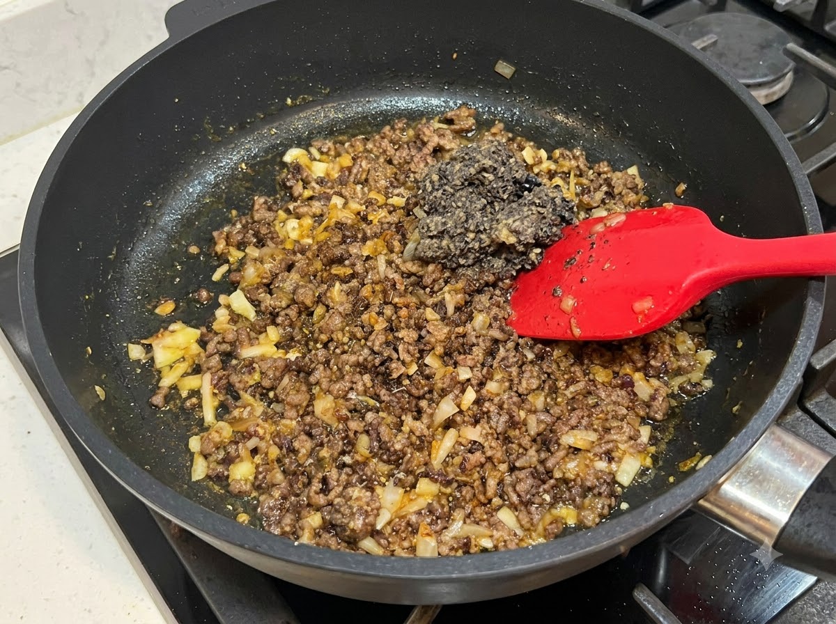 Ein roter Pfannenwender mischt einen dunklen Löffel pürierter schwarzer Bohnenpaste in eine Bratpfanne mit gekochtem Hackfleisch und Zwiebeln.