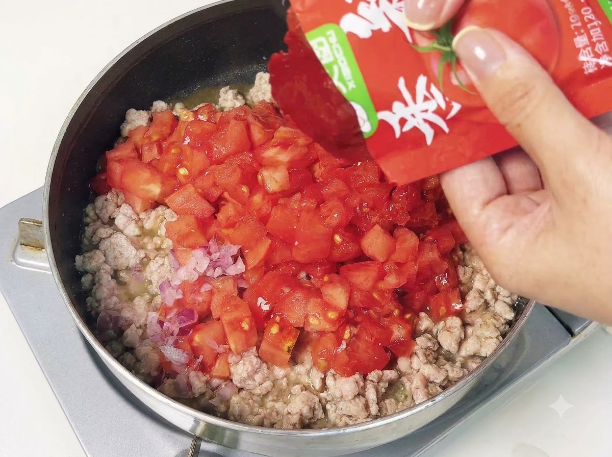 Eine Portion Tomatenmark wird in eine Pfanne mit Hackfleisch und gewürfelten Tomaten gegeben.