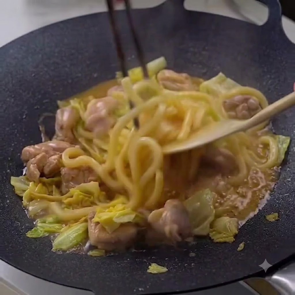 Essstäbchen und ein Holzspatel rühren dampfende Udon-Nudeln, Hähnchen und Kohl in einer dunklen Pfanne um.