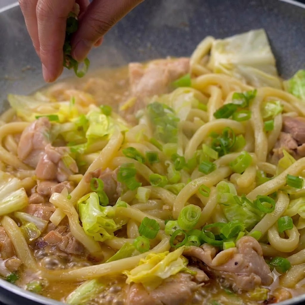 Eine Hand streut frisch gehackte Frühlingszwiebeln über eine Pfanne mit dampfenden Udon-Nudeln und Hähnchenstücken.